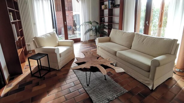 A living room with a sofa, armchair, and a small table. There is a bookshelf and a potted plant. - Spacious, family-friendly vacation apartment in architect's house with garden (Lauf)