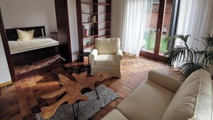 A modern living room with a sofa, a chair, a bed with a headboard, a wooden coffee table, a bookshelf, and a view of a brick building through a glass door. - Spacious, family-friendly vacation apartment in architect's house with garden (Lauf)