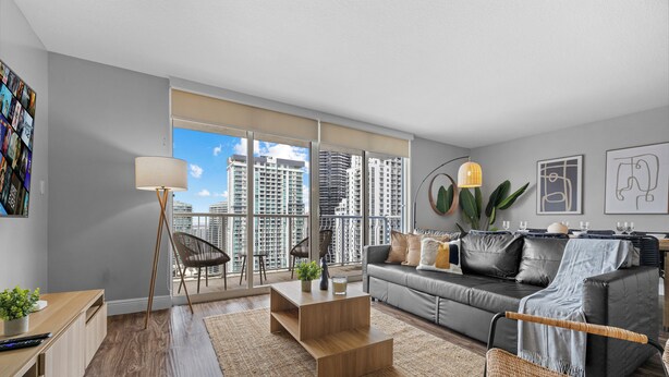A modern living room with a grey sofa, a coffee table, a floor lamp, and a view of high-rise buildings through large windows. - Chic Brickell Condos – Prime Location (Miami)