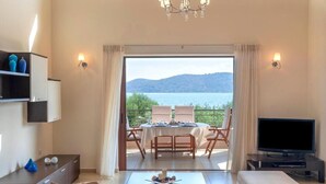 A living room with a TV, a dining table with chairs, and a view of a lake. - Elounda Blue Island Villa (Agios Nikolaos)