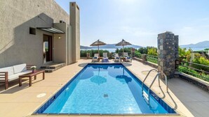 A rectangular swimming pool with a clear blue water surface, surrounded by lounge chairs and umbrellas, with a view of mountains in the background. - Elounda Blue Island Villa (Agios Nikolaos)