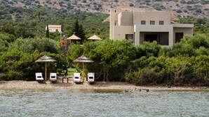 A modern building with a flat roof, surrounded by lush greenery and a body of water. There are lounge chairs and umbrellas on the shore. - Elounda Blue Island Villa (Agios Nikolaos)