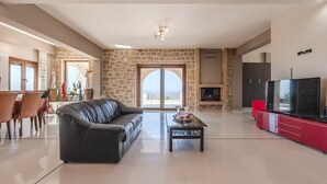 A spacious living room with a stone wall, a leather sofa, a glass coffee table, a red entertainment unit, and a large TV. - Arios Lux villa,50 sqm pool,Rural,8 guests,Rethymno (Agia Paraskevi)
