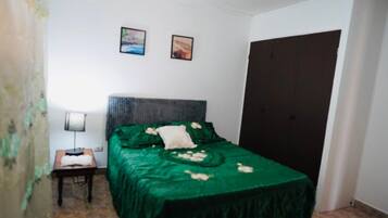 Een kleine, eenvoudige kamer met een bed, nachtkastje, lamp en een deur.