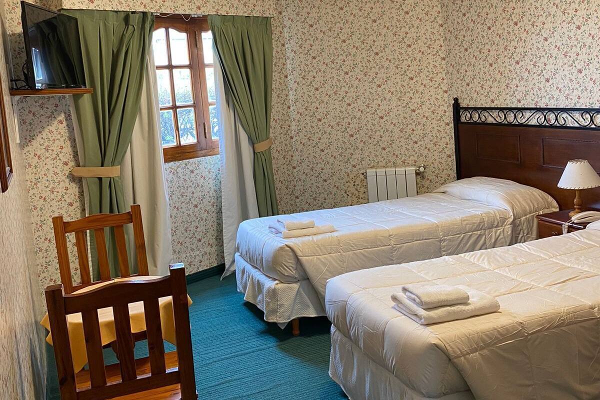 A room with two beds, a wooden chair, a television on a shelf, and a window with curtains.