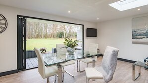 Un comedor moderno con una mesa y sillas de vidrio, un ventanal con vistas a un jardín y un televisor montado en la pared.