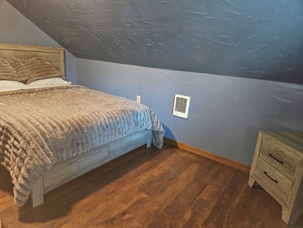 A bedroom with a bed, a nightstand, a wall vent, and wooden flooring.