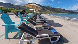 Een strand met rijen ligbedden en parasols, een hut met rieten dak en bergen op de achtergrond.