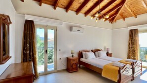 Une chambre avec un plafond en bois, un grand lit, une table de chevet, une commode et une porte-fenêtre coulissante donnant sur un balcon.