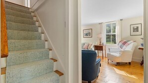 A staircase with a patterned carpet, a blue armchair, a white armchair, and a window with a view of greenery. - Beauchamp Cottage, near Rockport Harbor and golf c (Rockport)