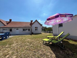 Een huis met een rode pannendak, een witte gevel en een kleine voortuin. Er staat een paarse parasol met 'HARIBO chamallows'-branding, een blauwe tafel met stoelen en twee groene loungestoelen.