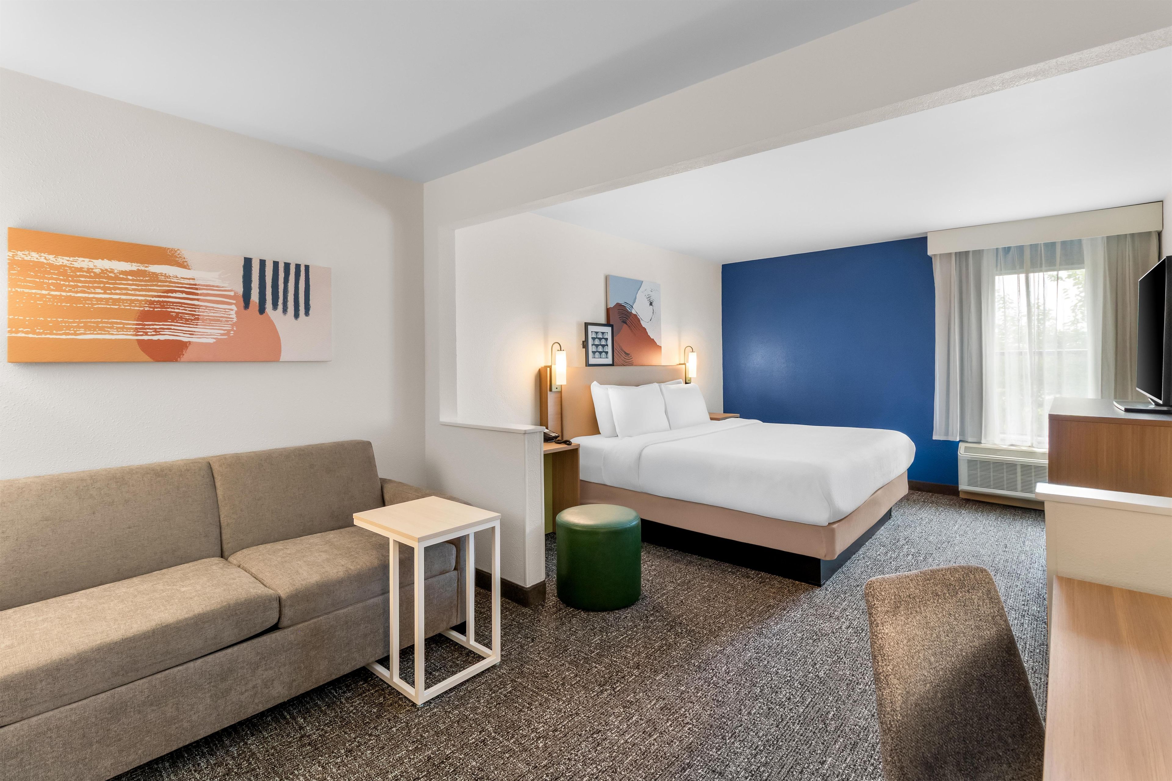 Ein Hotelzimmer mit einem Bett, einer Couch, einem kleinen Tisch, einem grünen Hocker und einem Fernseher.