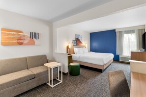 A hotel room with a bed, a sofa, a small table, a green stool, and a TV. - Spark by Hilton Dayton Fairborn (Fairborn)