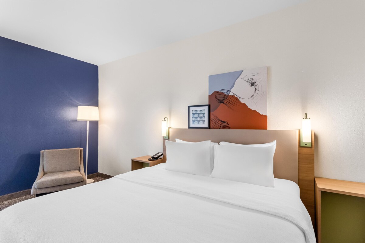 Una habitación de hotel con una cama, una silla, una mesita de noche con un teléfono y un cuadro en la pared.