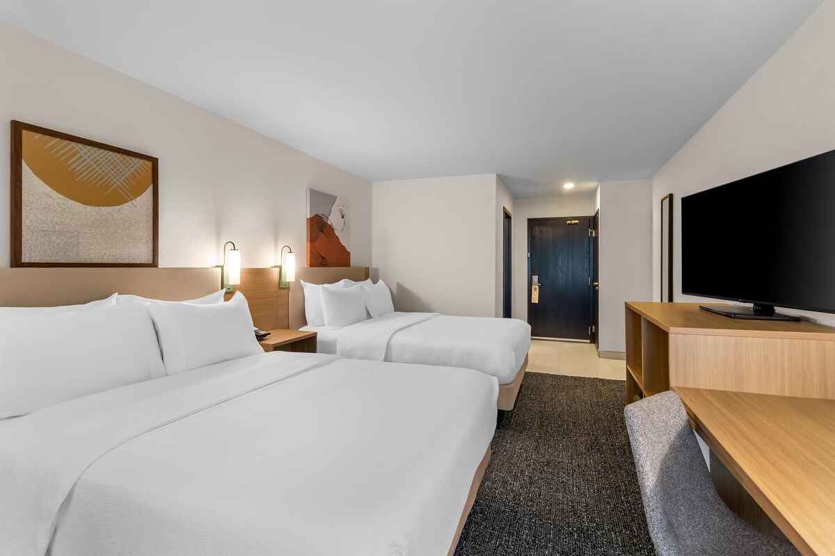 Una habitación de hotel con dos camas, televisión, escritorio y obras de arte en las paredes.