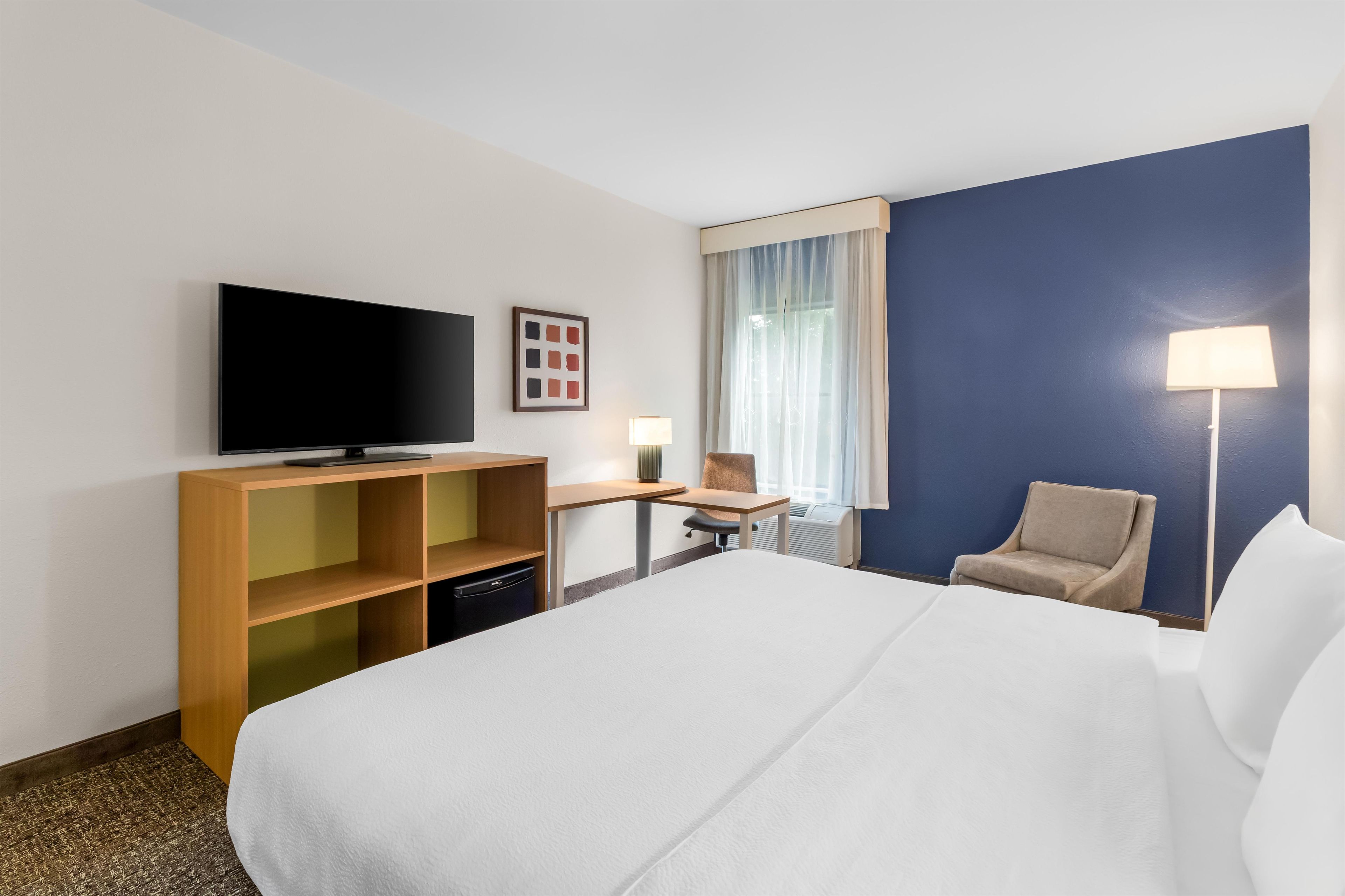 Habitación de hotel con cama, escritorio, silla, televisión y ventana con cortinas.