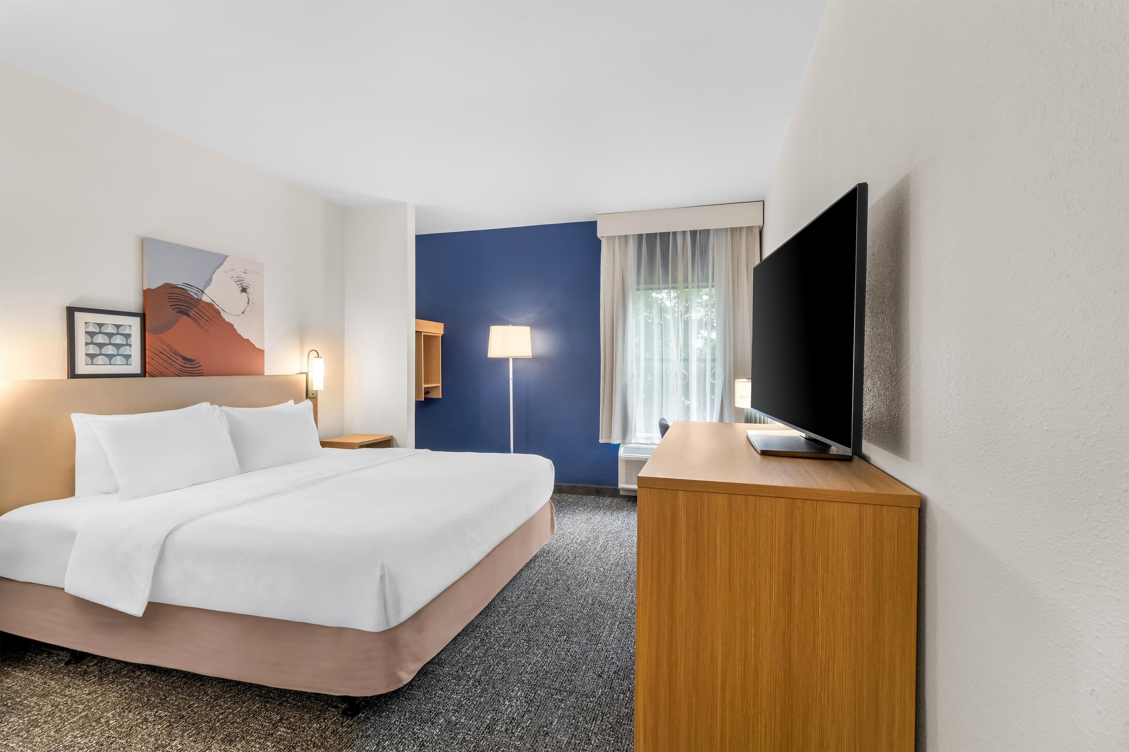 A hotel room with a bed, a flat-screen TV, a desk, a lamp, and a painting on the wall.
