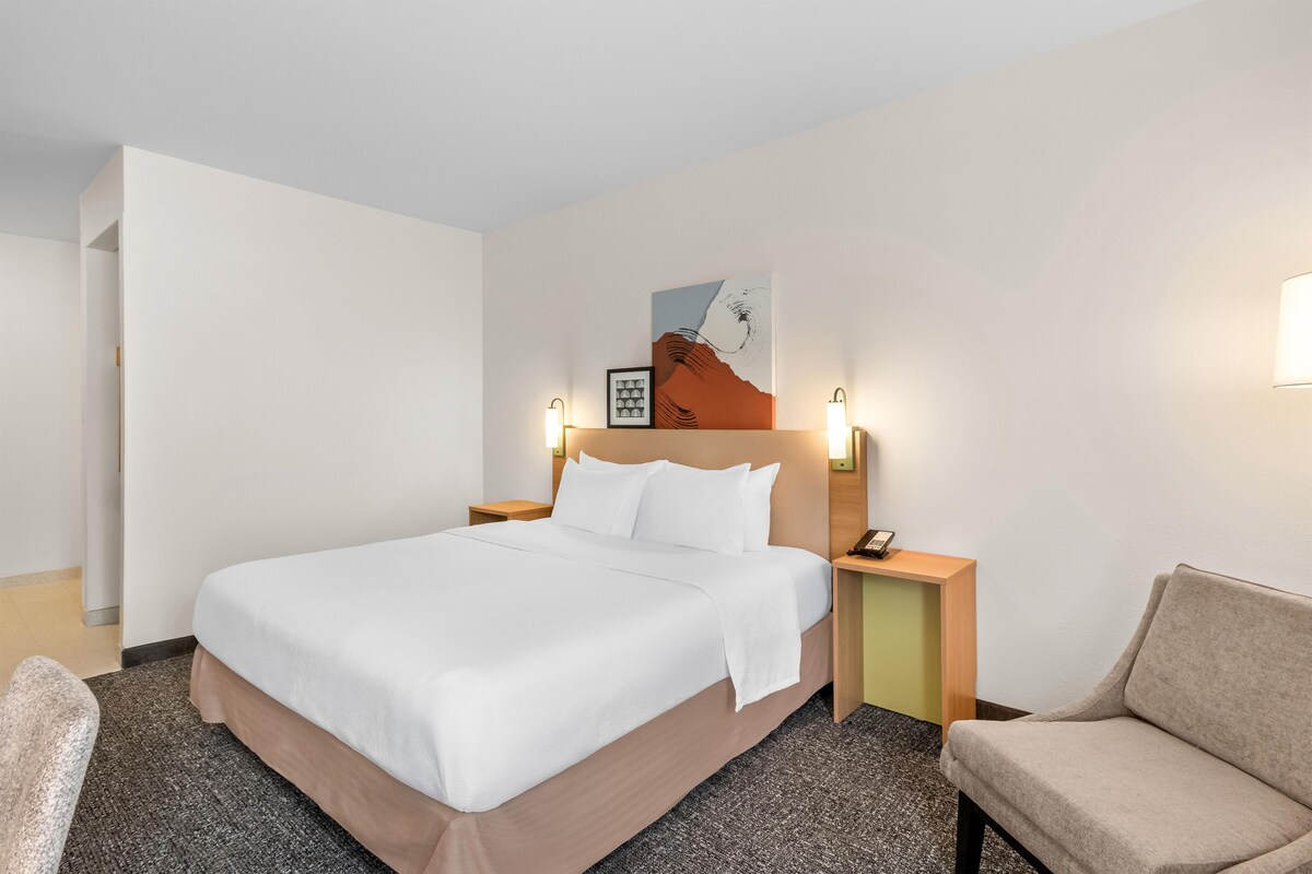 Una habitación de hotel con cama, mesitas de noche, una silla y un cuadro en la pared.