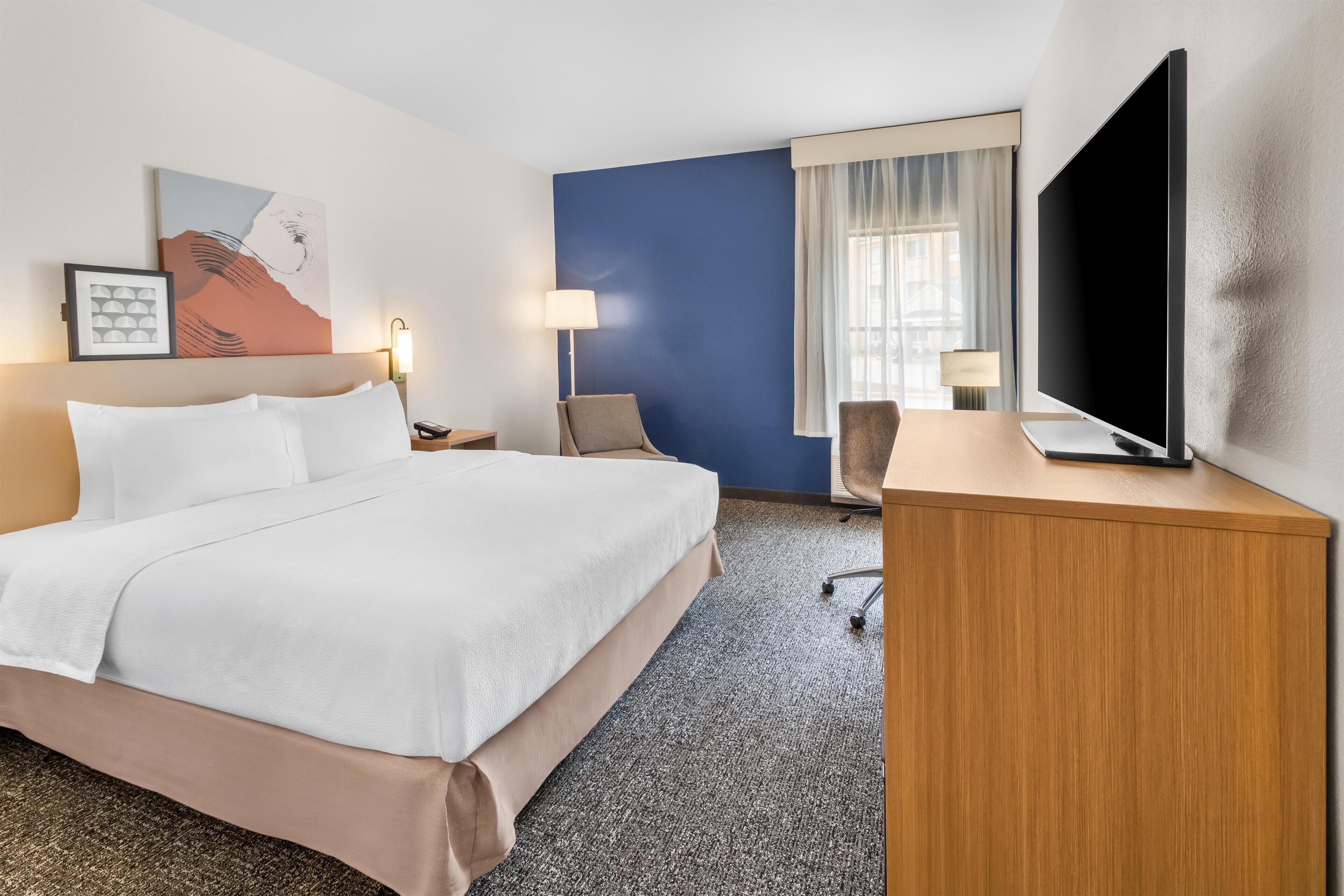 Habitación de hotel con cama, escritorio, silla, televisor y ventana con cortinas.