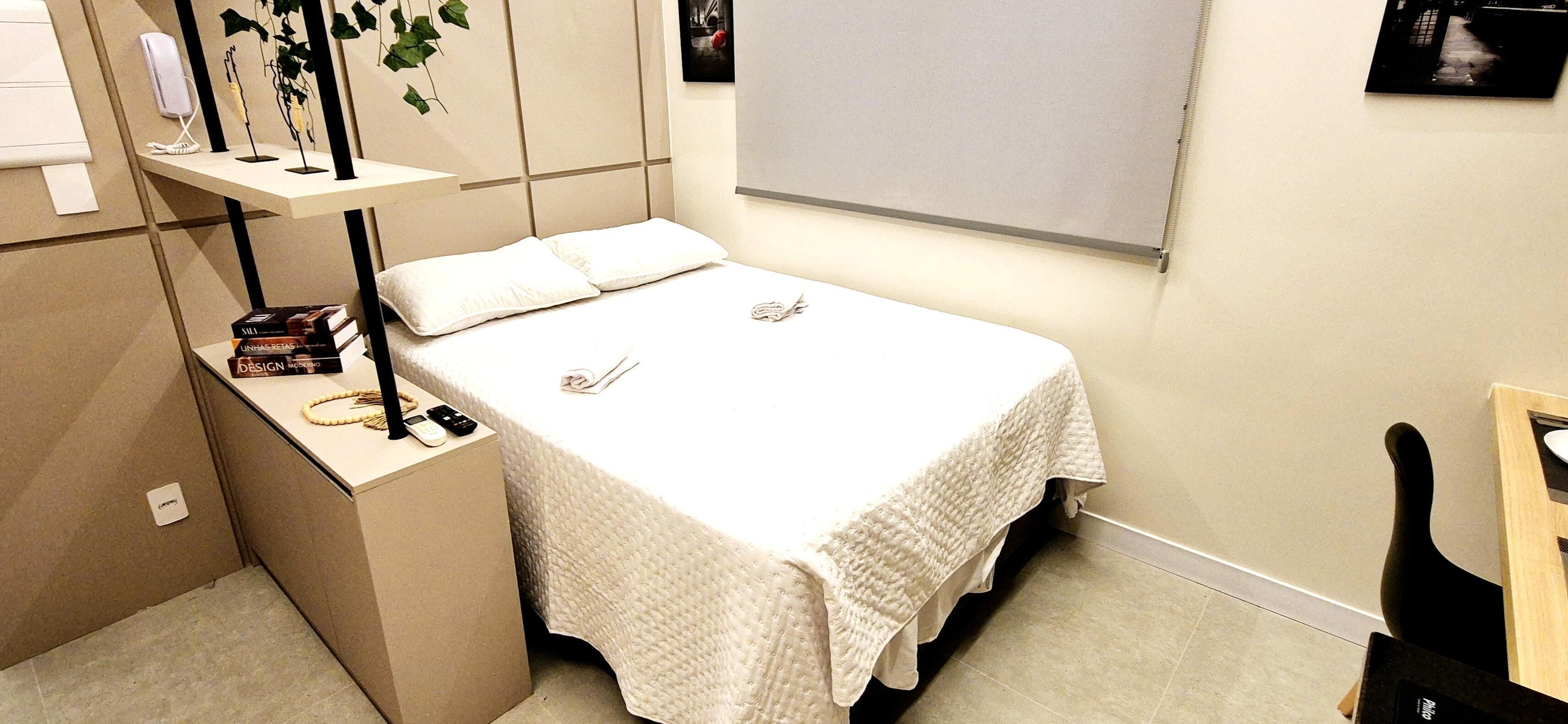 Une chambre à coucher bien rangée, avec un lit, une table de chevet et une petite étagère contenant des livres et un téléphone.