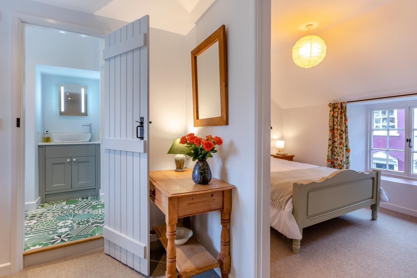 A bedroom with a bed, a bedside table with a vase of flowers, a nightstand, and a window with curtains.