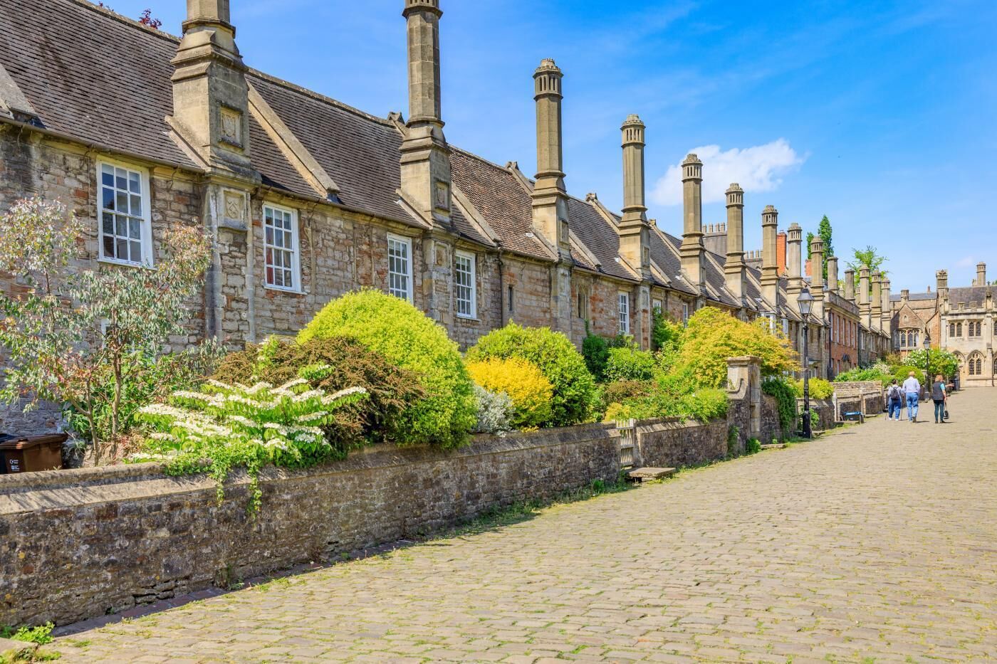 A row of stone buildings with multiple chimneys, a cobblestone street, and a stone wall with greenery.