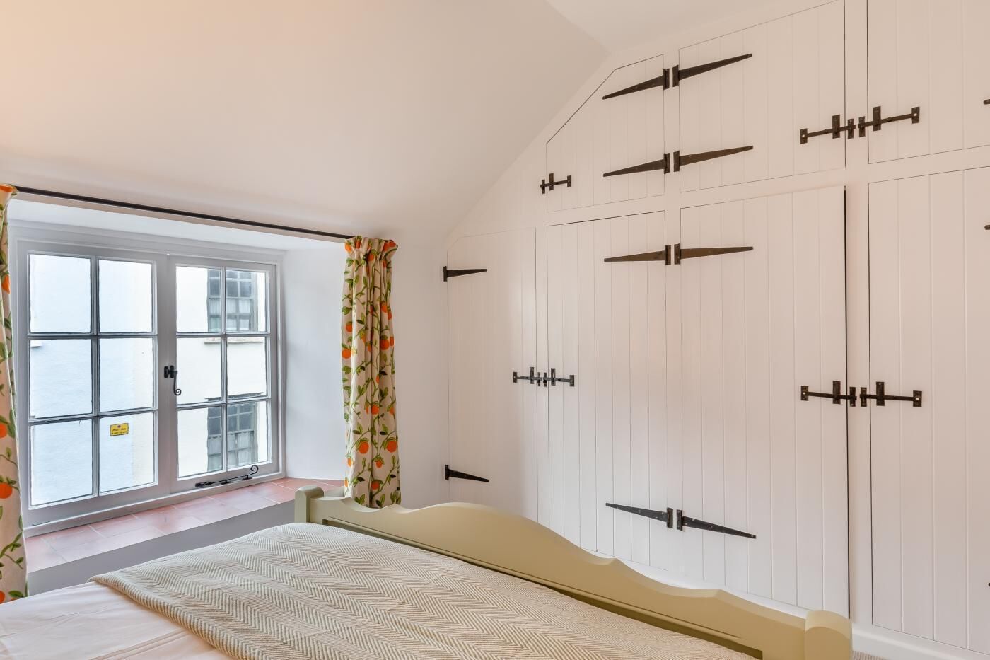 A bedroom with a large window, a bed with a patterned bedspread, and a wardrobe with multiple doors.