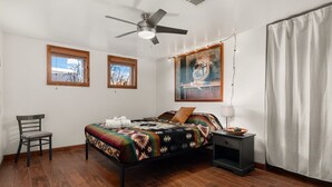 A bedroom with a bed, a chair, a nightstand, a ceiling fan, and two windows. - Native Charm Adobe Guesthouse in Historic Old Town with wifi, AC, patio grill. (albuquerque)