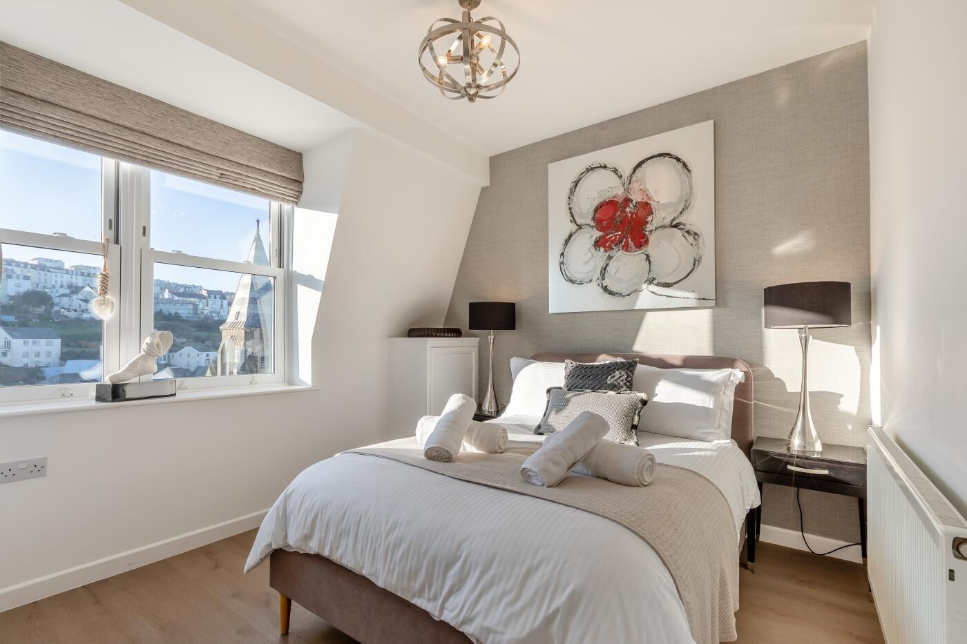 Une chambre à coucher moderne, dotée d’un grand lit, offrant une vue sur le paysage urbain et agrémentée d’un tableau abstrait original au mur.