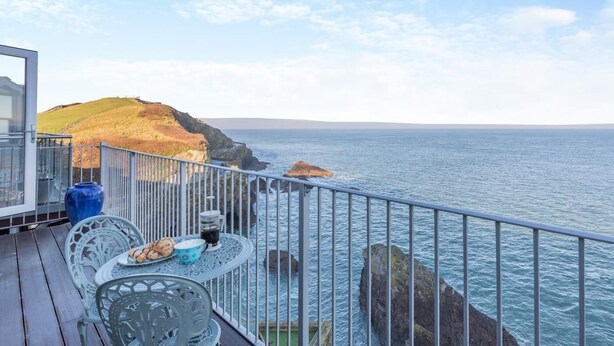 A deck with a table and chairs overlooking a coastal view with rocky cliffs and the sea. - 2 Bed in Ilfracombe (oc-o30532) (Ilfracombe)