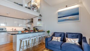 A modern kitchen with white cabinets, a central island with bar stools, and a blue sofa in the living area. - 2 Bed in Ilfracombe (oc-o30532) (Ilfracombe)