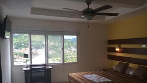 A bedroom with a bed, a desk, and a chair. There is a window with a view of a cityscape and mountains. - Hotel Tijerinos (Boaco)