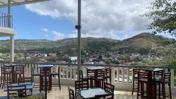 An outdoor terrace with tables and chairs, overlooking a hilly landscape with buildings. - Hotel Tijerinos (Boaco)