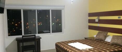 Un dormitorio con cama, escritorio y silla. Se ve una ventana con vistas a luces de la ciudad.