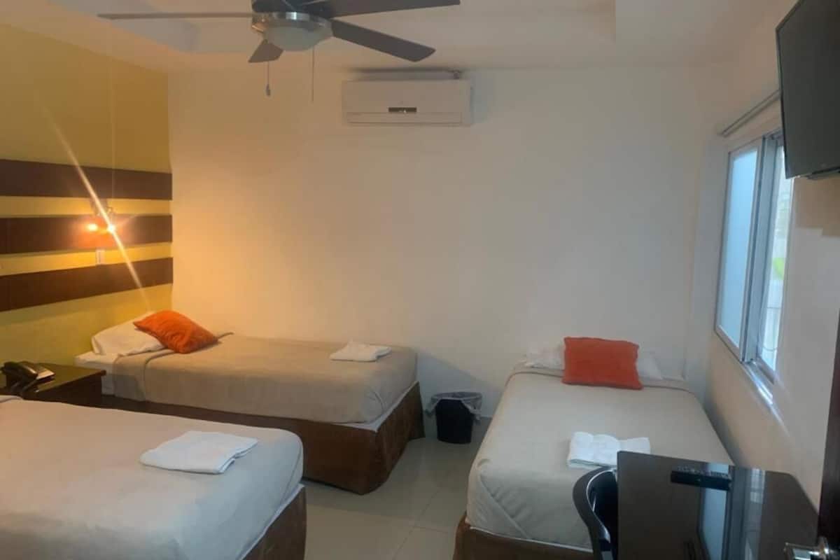 A hotel room with two single beds, a ceiling fan, an air conditioning unit, a desk with a chair, and a window providing natural light.