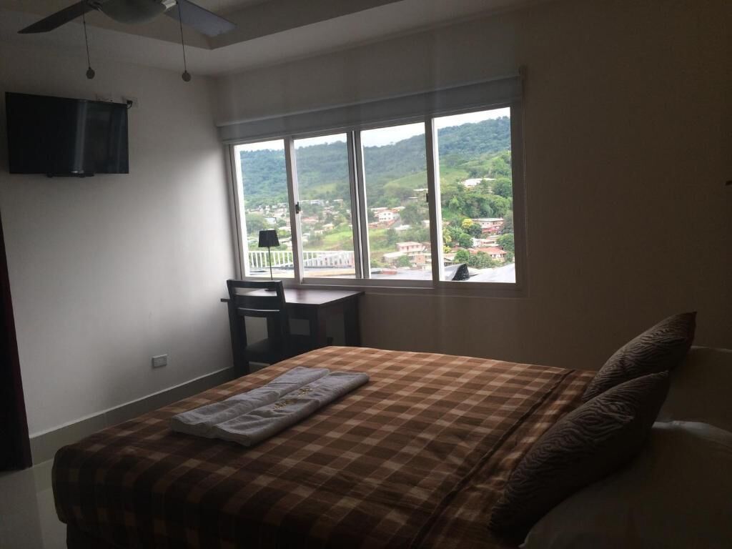 A bedroom with a bed, a desk, and a large window offering a view of a town and hills.