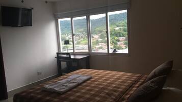 A bedroom with a bed, a desk, and a large window offering a view of a town and hills.