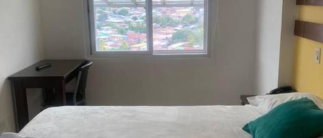 A hotel room with a bed, a desk, and a window with a view of buildings.