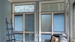 Ein modernes Wohnzimmer mit einem weißen Sofa, einem schwarzen Couchtisch und einem gemusterten Teppich. Große Fenster mit Jalousien, ein Deckenventilator und eine Wandleiter sind ebenfalls vorhanden.