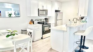 A modern kitchen with white cabinetry, a microwave, a refrigerator, a stove, and a dining area with white chairs and a table. - Stunning Family Getaway - One Block from Beach, Fast WiFi, A/C, BBQ, Parking (San Diego)