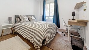 Une chambre à coucher comprenant un lit, un bureau, une chaise, une petite étagère, une corbeille à déchets et une fenêtre avec des rideaux.