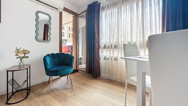 Un salon moderne avec un fauteuil bleu, un bureau blanc et une grande fenêtre ornée de rideaux légers.