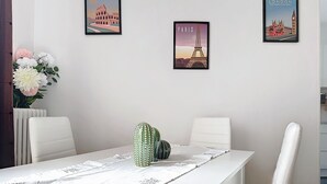 Un coin repas avec une table blanche, deux chaises blanches, un cactus et trois tableaux représentant des monuments célèbres.