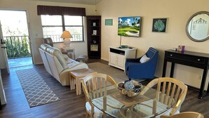 Ein Wohnzimmer mit einem Esstisch mit Glasplatte, Holzstühlen, einem blauen Sessel, einer Couch, einer kleinen Kommode und einem großen Spiegel an der Wand.