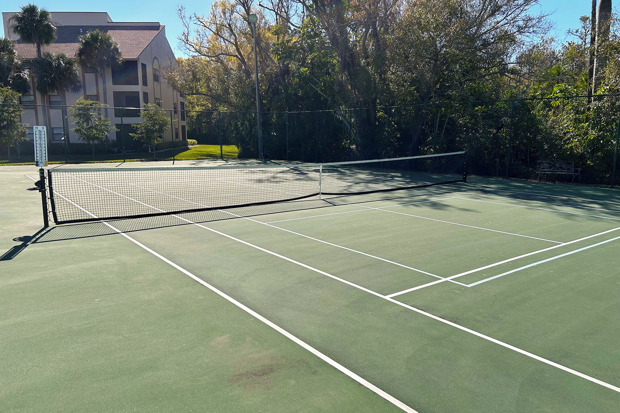 A tennis court with a net, surrounded by trees and a building in the background.