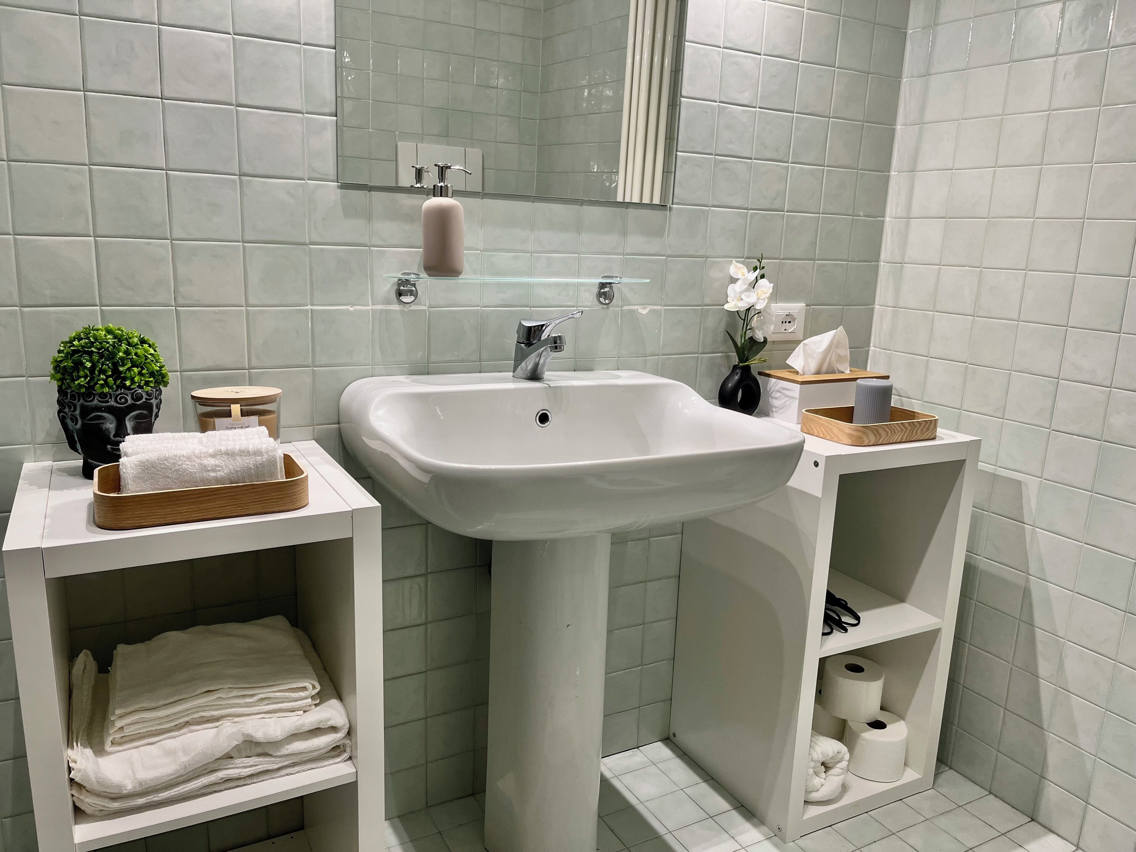 A modern bathroom with a white sink, a mirror, and a toilet. There are shelves with towels and toiletries.