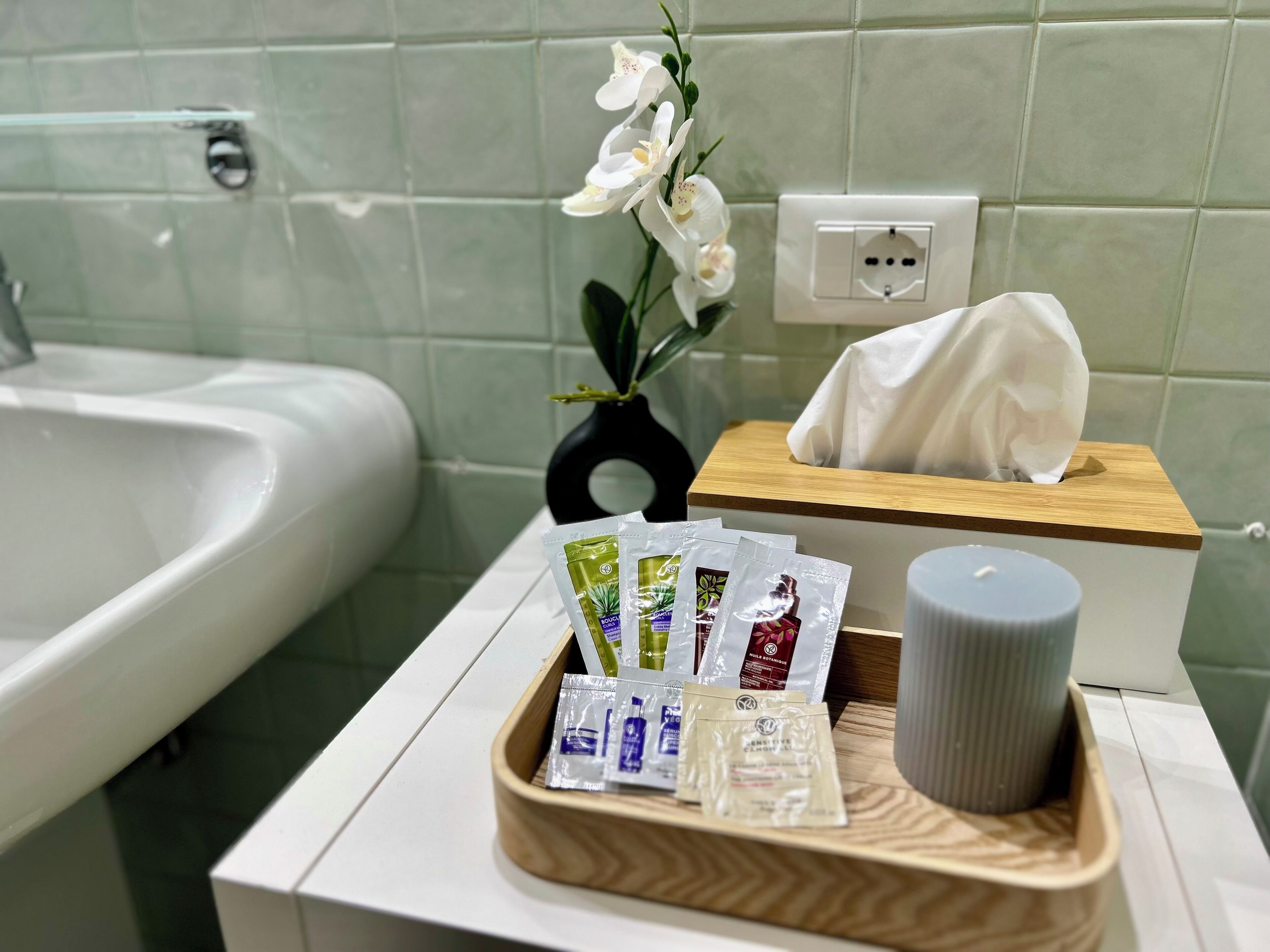 Un bagno con un lavandino bianco, un vassoio di legno con articoli da toilette, un portascocche e un vaso con fiori bianchi.