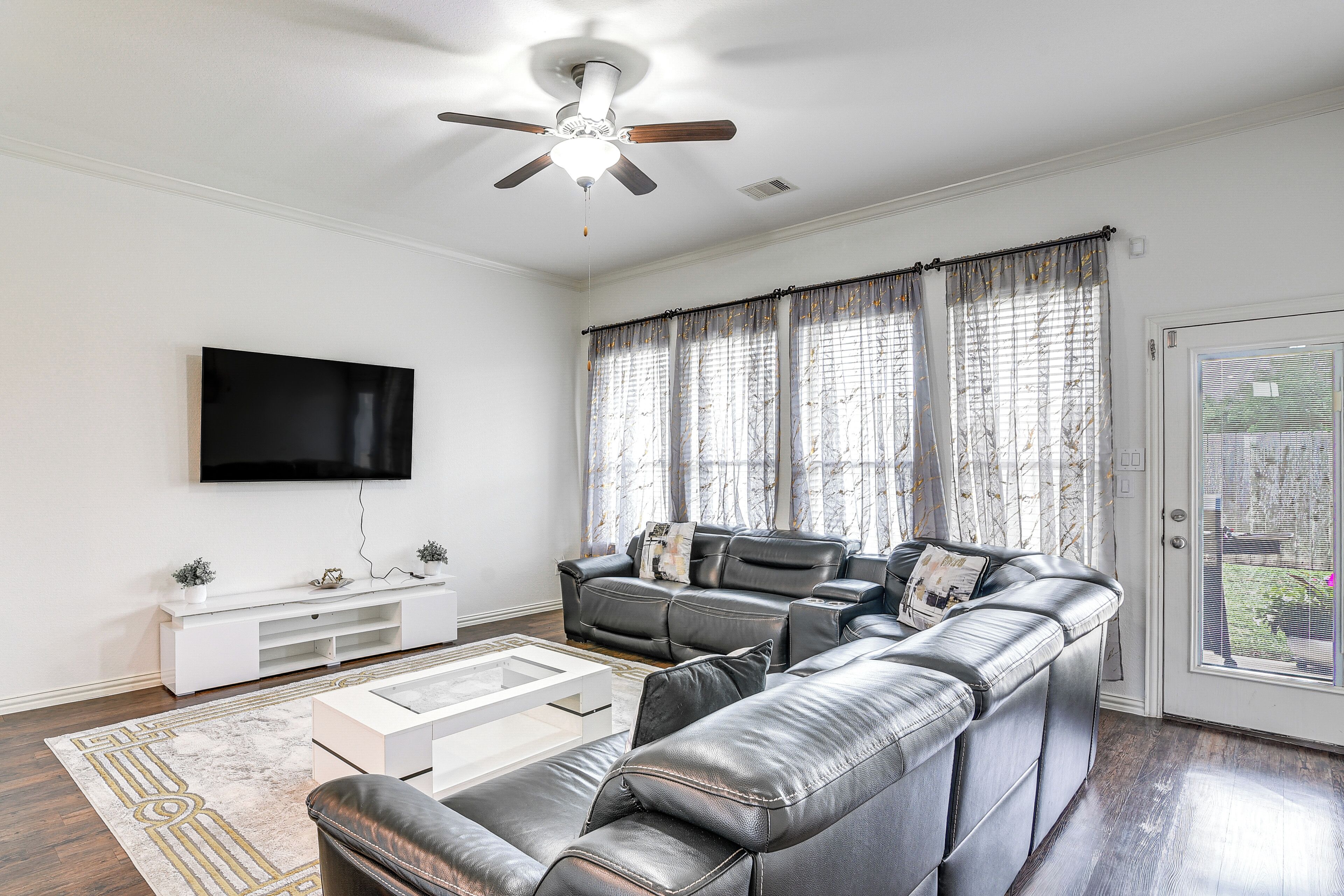 Ein Wohnzimmer mit Ecksofa, Flachbildfernseher, Deckenventilator und großen Fenstern mit Vorhängen.