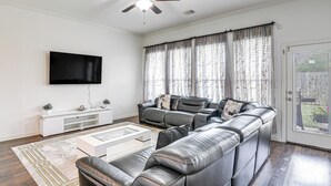 Una sala de estar con un sofá modular, un televisor de pantalla plana, un ventilador de techo y ventanas grandes con cortinas.
