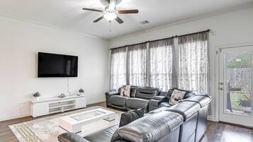 Ein Wohnzimmer mit Ecksofa, Flachbildfernseher, Deckenventilator und großen Fenstern mit Vorhängen.